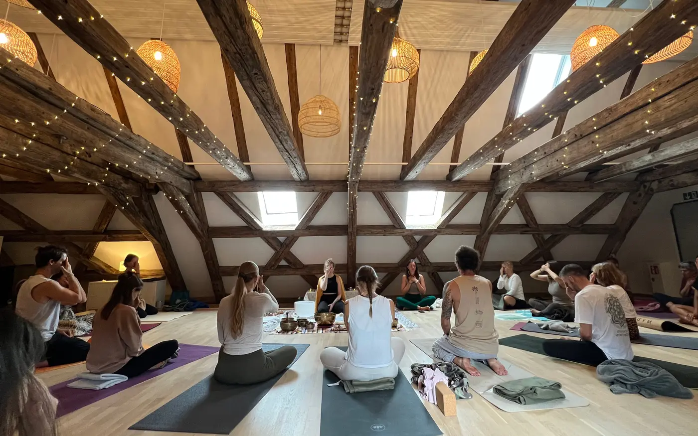Ein Gruppen-Klangbad beim Bhakti Summer Yoga Festival, September 2026: die Teilnehmenden sitzen auf ihren Matten unter alten Holzbalken, Anna zugewandt